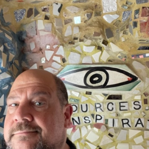 Frank Pellicone smiling in front of a wall filled with mosaic pieces and a large eye.