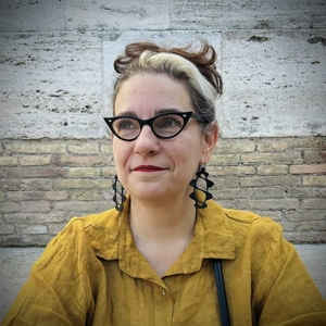 Arielle Saiber wearing yellow shirt, large black earrings with helices, and black cat-eyed glasses standing in front of a brick building