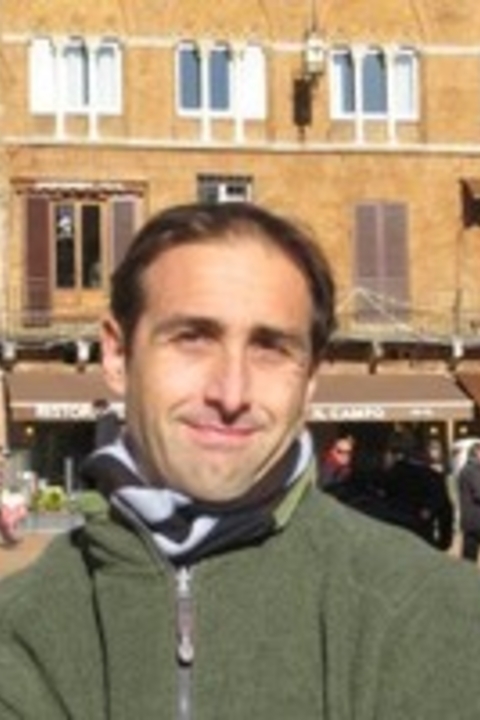Enrico Cesaretti with short brown hair, grey and black scarf, green jacket standing in Siena's Piazza del Campo.