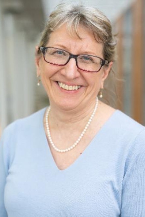 A photograph of Marilyn Migiel with hair pulled back, glasses, wearing a light blue v neck sweater, pearl necklace and earrings.