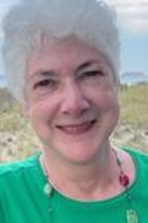 A photograph of Ann Mullaney wearing a green shirt, grey short hair with ocean in the background.