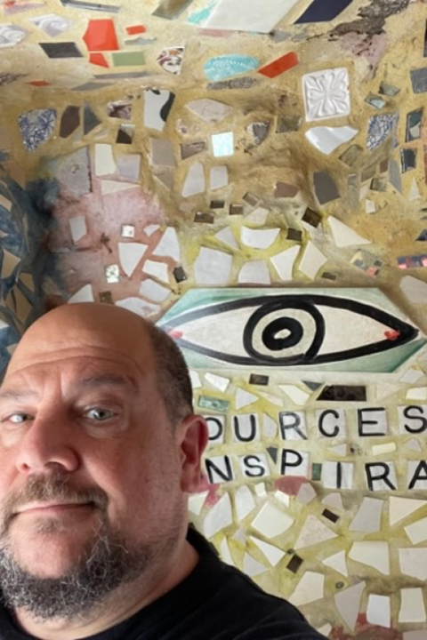 Frank Pellicone smiling in front of a wall filled with mosaic pieces and a large eye.