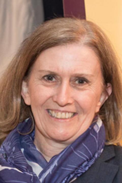 A photograph of Susan Briziarelli smiling at the camera, wearing a white scarf with large dark blue flowers on it.