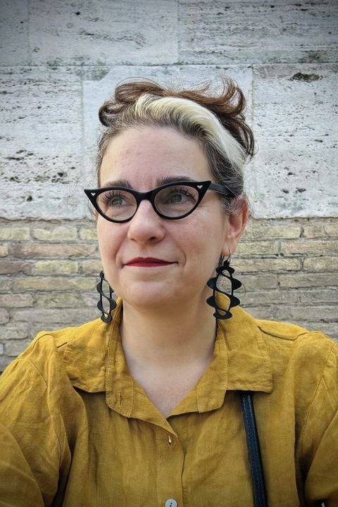 Arielle Saiber wearing yellow shirt, large black earrings with helices, and black cat-eyed glasses standing in front of a brick building