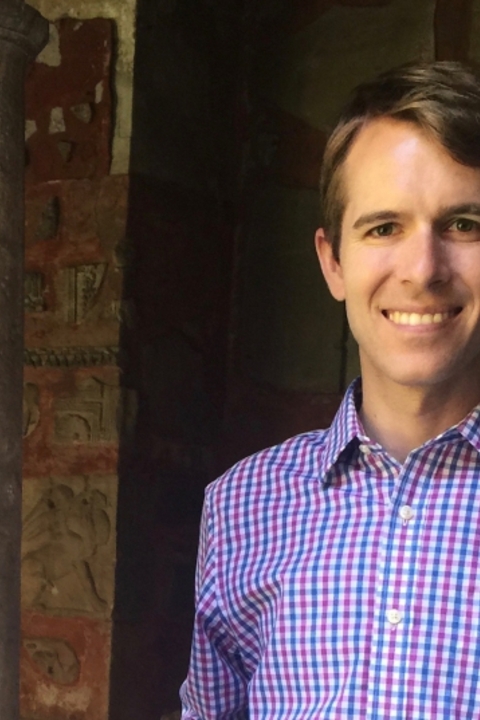 James Coleman wearing a blue and red plaid shirt standing in front of a old building