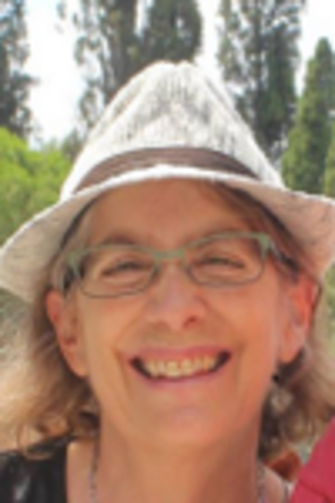 A photo Stephanie Jed, wearing a fedora, glasses, and smiling into the camera.